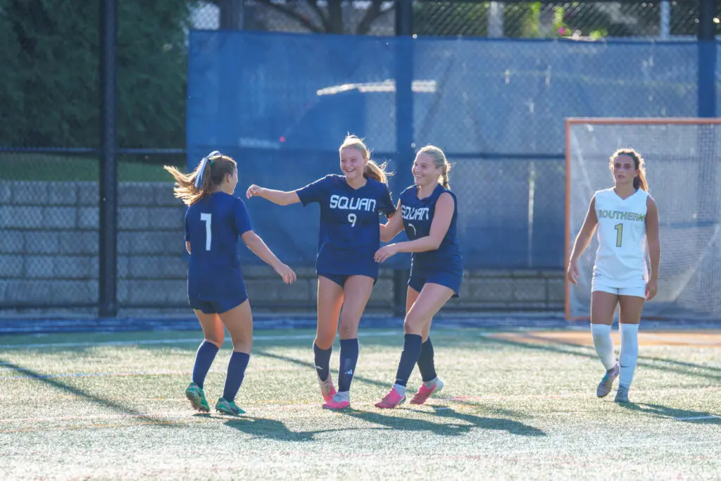 SSI_Girls_Soccer_Manasquan_v_Southern_9-11-26-53 - Shore Sports Insider Lexie Liebler (middle) celebrates with Brooke Mack (Left) and Madison Decker (right) after scoring against Southern. 9/11/26 Photo by Patrick Olivero - SSI_Girls_Soccer_Manasquan_v_Southern_9-11-26-53' soccer teammates in blue celebrate and smile on a sunny field near the goal net, after a play.