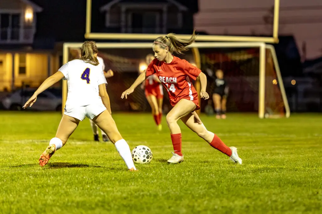 Two female soccer players chase a ball on a lit night field, one in red (#14) and one in white (#4) near the goal.