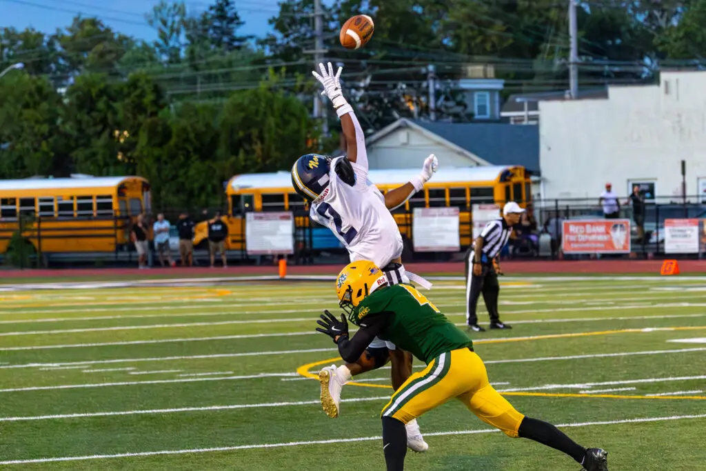 Football game action: a white-jersey player leaps to catch a high pass while a green-and-yellow defender reaches in front on a turf field with bleachers in the background.
